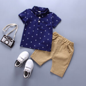 Flat lay of a toddler boy's summer outfit featuring a navy blue short-sleeve polo shirt with an all-over white anchor pattern, a pair of tan khaki shorts with an elastic waistband, small white velcro sneakers with dark accents, and a little fabric camera accessory.